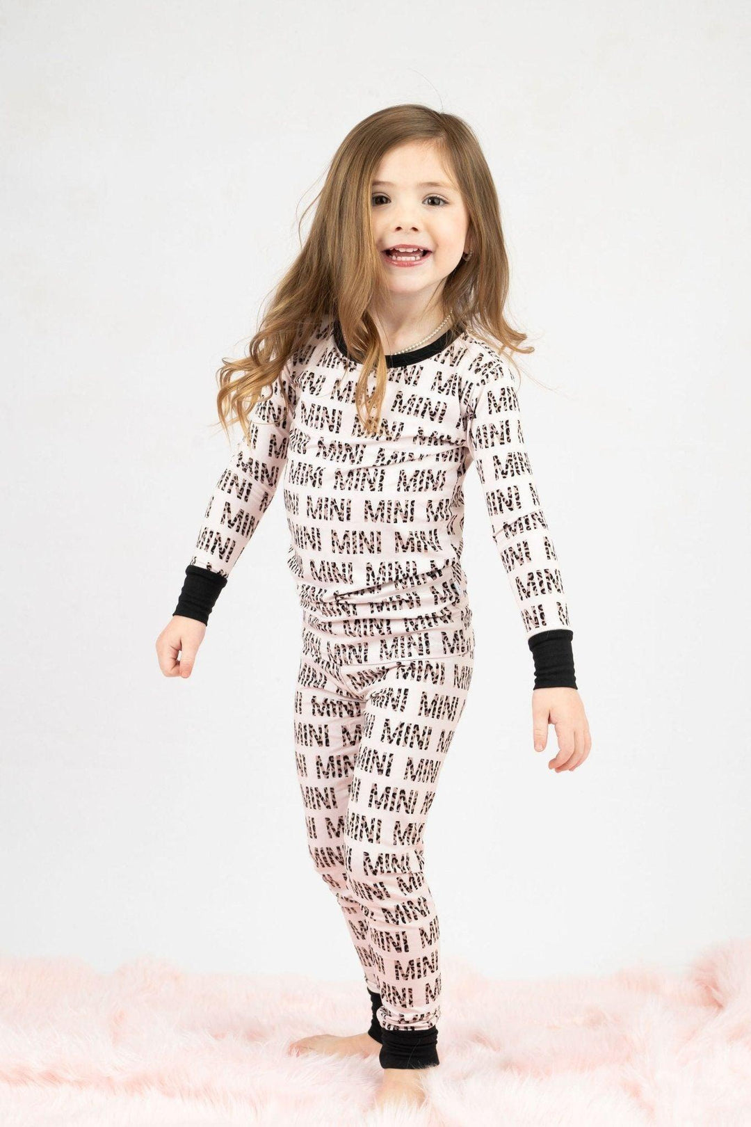 A young girl with long brown hair, smiling and walking towards the camera, wearing our bamboo two-piece pajama set with the words "mini" written in leopard print, against a white background.